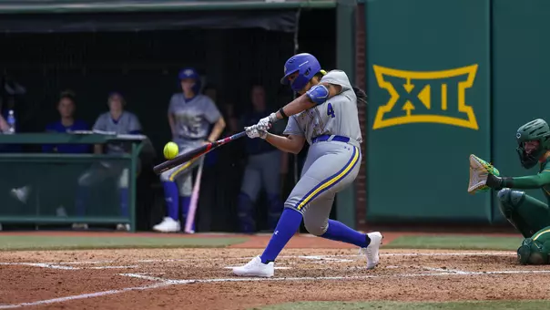 A woman in a grey uniform swings a softball bat and hits a softball into the air