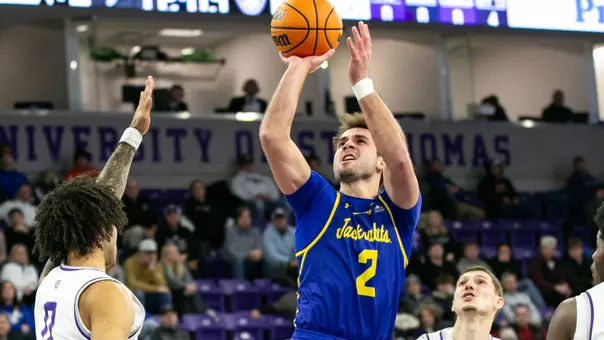 A men's basketball player in a blue uniform shoots the basketball over two defenders in white uniforms