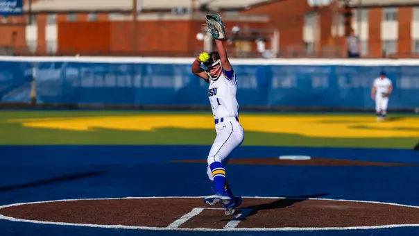 A women's softball player, Amanda Vacanti, in a white uniform winds up to a throw a pitch towards home plate