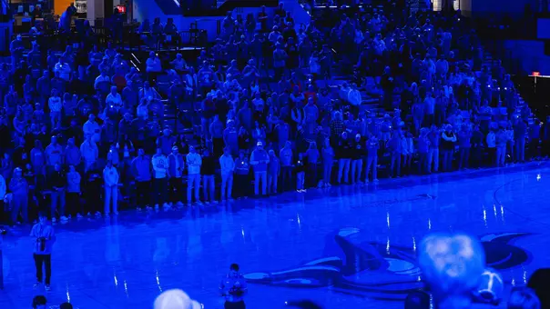 Blue light shines down over fans clapping in the seating section and a basketball court prior to a basketball game
