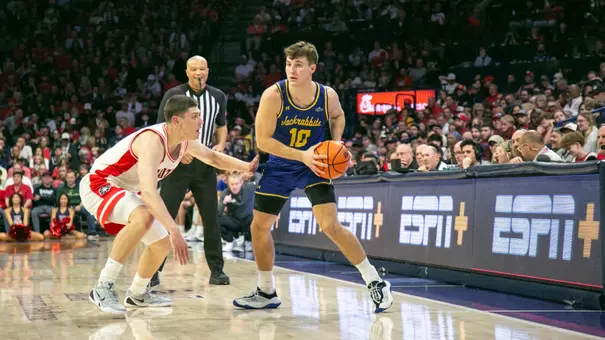 A men's basketball player, Kalen Garry, in a blue uniform holds a basketball on a court while being defended by a player in a white uniform