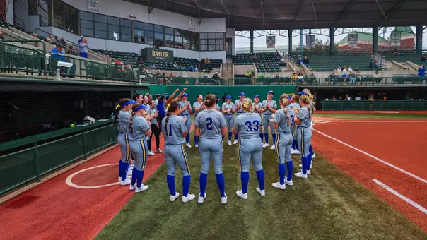 A softball team in white uniforms stands in a huddle on the side of a softball field prior to a game