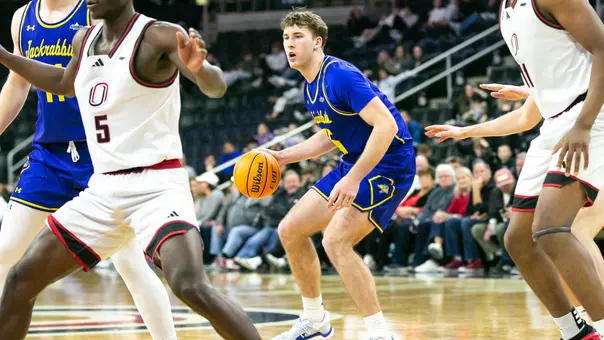 A men's basketball player in a blue uniform, Alec Squires, holds a basketball in his right hand while surveying the court