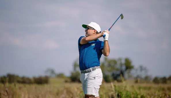 A male golfer, Bryson Mercier, in a blue patterned polo and white cap follows through on his swing on a golf course under a clear sky.