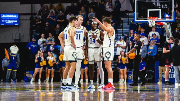 A group of men's basketball players in white uniforms huddle at center court during a game, with cheerleaders and spectators visible in the background. A referee holding a basketball stands nearby, and the scoreboard overhead shows 25 seconds on the shot clock.