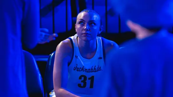 A women's basketball player in a white uniform sits on the bench during pregame lineup introductions