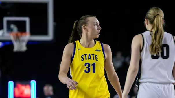 A women's basketball player in a yellow uniform walks up the court during a game. A player from the opposing team in a white uniform is also visible in frame.