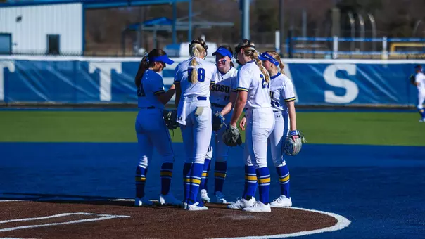 A group of softball players in white and blue uniforms stands together in a huddle on the pitching mound during a game on a blue and brown turf field. One player stands off to the side near the outfield fence. Stadium fencing, dugouts, and trees are visible in the background.