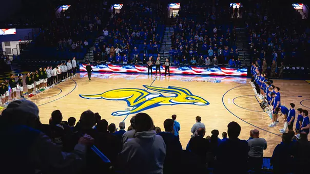 A basketball arena filled with spectators stands for a pregame ceremony. Two teams line up on opposite sidelines of the court, which displays a large yellow-and-blue jackrabbit logo at center. A long digital board along the scorer’s table shows an American flag graphic.