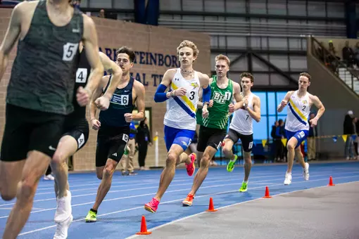 A male track runner in white races around the curve of the track surrounded by competitors jockeying for position