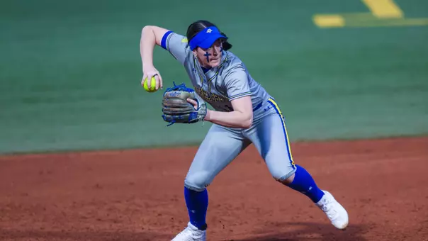A woman in a grey uniform with blue details prepares to a throw a softball across the diamond