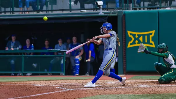 A woman in a grey uniform with blue details swings a softball bat and makes contact with the ball going into the air