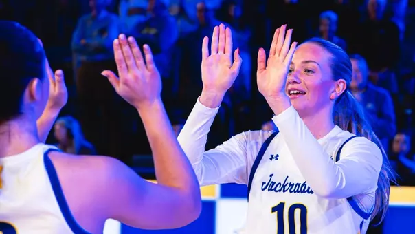 Women's basketball play in a white uniform goes through a high five line with teammates during pregame lineup announcements. The arena is lit with blue light.