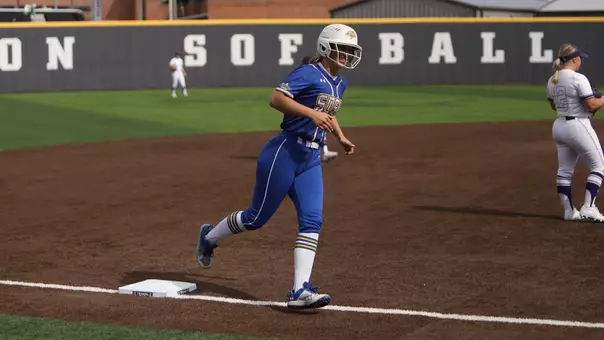 A woman in a blue softball uniform with a white helmet rounds third base and is smiling while running on a softball field