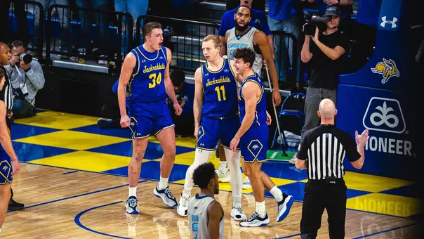 A college basketball game scene shows players gathered under the basket after a play. Three players in blue and yellow uniforms stand near the hoop, while an official gestures near them. A player in a light-colored uniform walks away from the basket. The court is surrounded by a crowded arena with fans in blue and yellow, and a scoreboard above the hoop displays 8:28 and a shot clock reading 30.