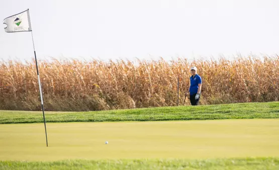 A women golfer in a blue polo and white hat stands next to a golf green observing her ball after her shot.