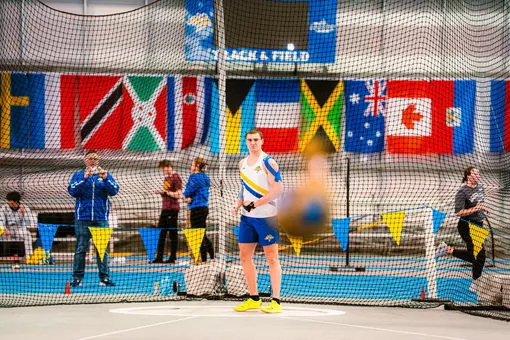 A male thrower watches his attempt land. The weight he threw is blurry and in front of the athlete