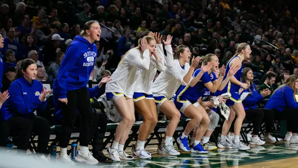 The South Dakota State bench celebrates during a game, with several players in white warmup shirts and blue uniforms standing and clapping.