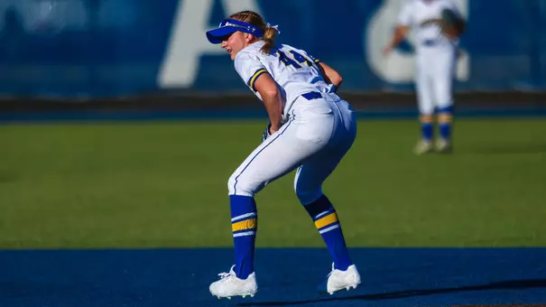A woman in a white softball uniform, with blue and yellow accents, is crouched over while preparing to throw a softball