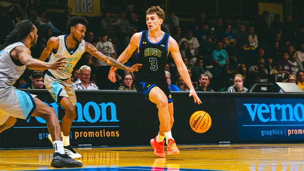 A man in a black uniforms, with blue and yellow accents, dribbles a basketball on a basketball court with defenders in grey uniforms surrounding him