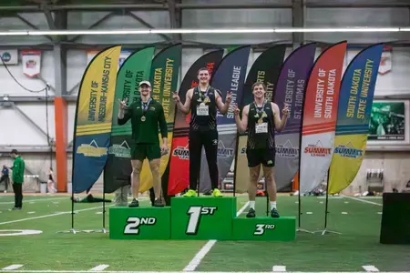 Three male weight throwers stand on the podium in front of school flags. The first and third-place finishers wear black, while the runner-up finisher is in dark green.