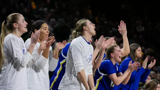 Members of the South Dakota State women's basketball team stand, clap, and cheer from the bench during a basketball game.