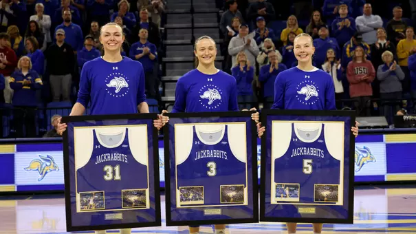 South Dakota State's three women's basketball seniors stand together holding framed jerseys in the middle of a basketball court. A full arena of fans is visible behind them.