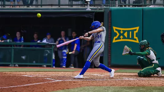 A woman in a grey softball uniform swings a softball bat and connects with the ball sending it into the air