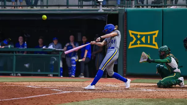 A woman in a grey softball uniform swings a softball bat and connects with the ball sending it into the air