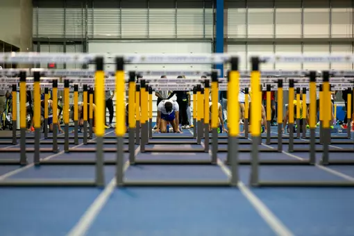 A male athlete is depicted in the starting blocks, shot through the middle of several hurdles on the 60-meter front stretch of the track