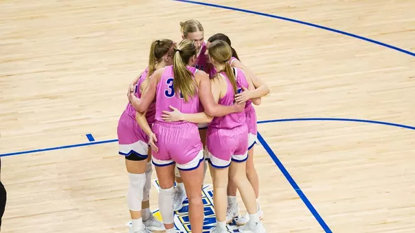 Women's basketball players in pink uniforms huddle on the court
