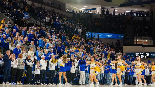 Cheerleaders in white uniforms on the court while fans in the stands in yellow and blue attire applaud