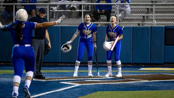 Two softball players in blue uniforms yell at home while watching a teammate run