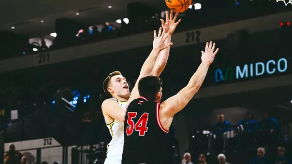 A men's basketball player in a white uniform shoots a shot with his left hand as a man in a black uniform defends him