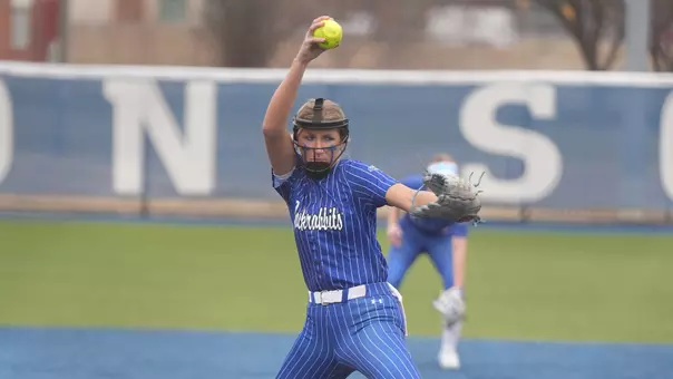 A softball pitcher in a blue uniform with white pin stripes begins to throw the ball from above her head towards the plate