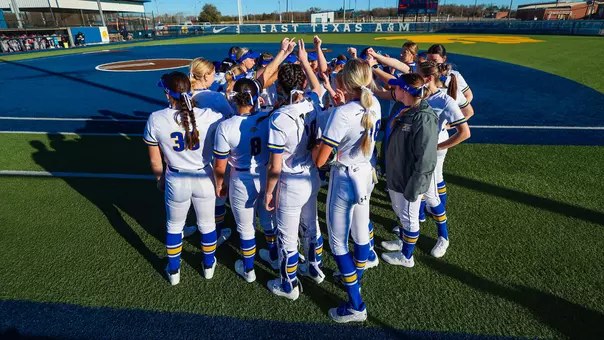A group of women in white softball uniforms put their hands up in a huddle on the side of a softball field