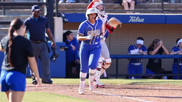 A woman in a blue softball uniform runs up the first base line of a softball field. A catcher in a white uniform behind the other woman throws a softball.