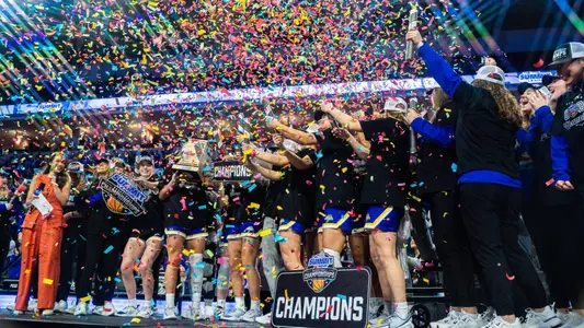 A women's basketball team celebrates a championship on stage on a basketball court as confetti falls around them
