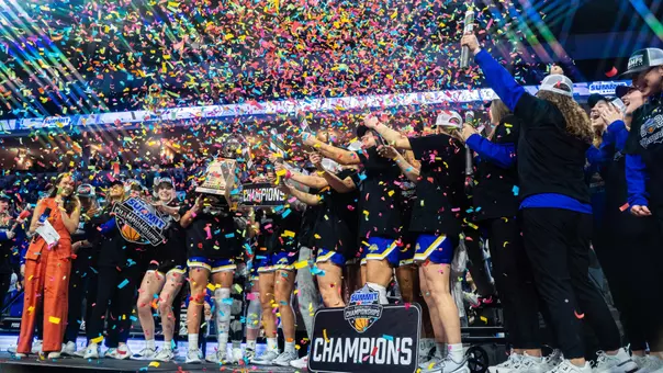 A women's basketball team celebrates a championship on stage on a basketball court as confetti falls around them