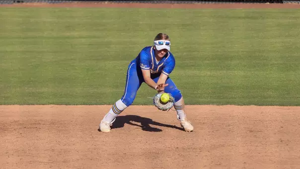 A woman in a blue softball uniform is crouched down to field a softball that bounces towards her while on a dirt infield