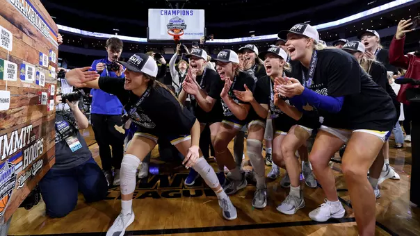 A women's basketball team wearing championship hats and t-shirts cheers as one teammate places a bracket sticker on the Summit League Tournament bracket