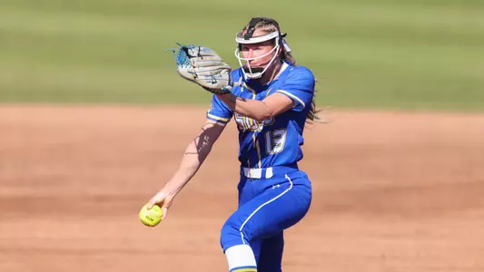 A woman in a blue softball uniform gathers momentum towards home plate while winding up to throw a pitch
