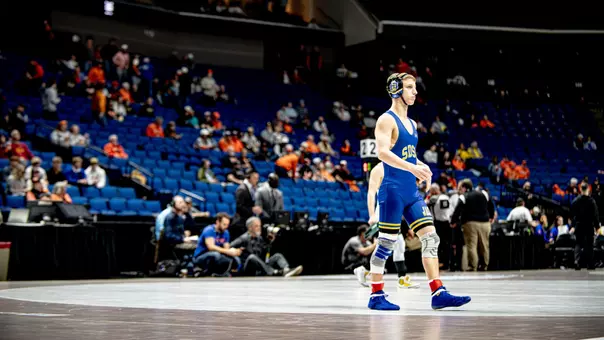 A wrestler in a blue singlet paces back in forth on a mat before a match.
