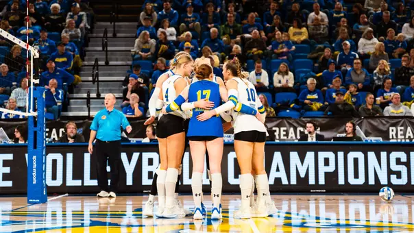 Women's volleyball players huddle on a court during a game. Fans in the stands are visible behind them.