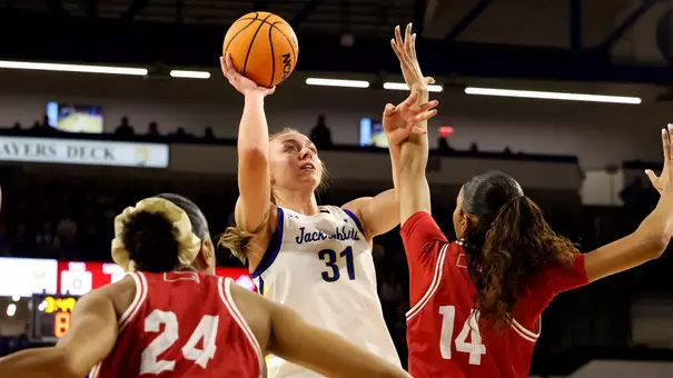 A women's basketball player in a white “Jackrabbits” jersey jumping with the ball extended overhead during a shot attempt against defenders in red uniforms.