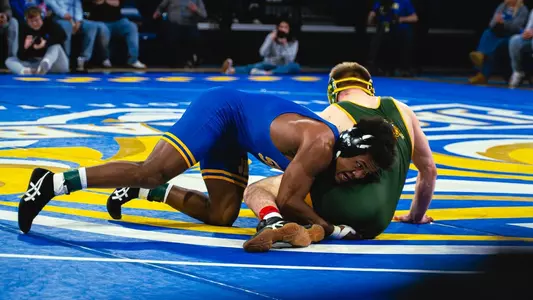 Marcus Espinoza-Owens, a wrestler in a blue singlet scores a takedown on a wrestler in a green and yellow singlet