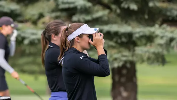A women's golfer wearing a white hat and black polo looks through a rangefinder on the golf course.