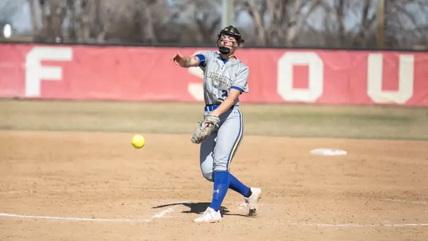 A woman in a grey softball uniform throws a softball towards home plate while pitching