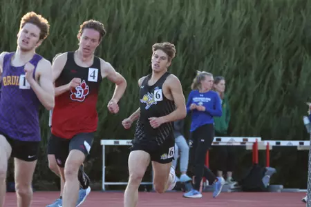 A male runner in black rounds the curve of the track in a long distance race behind two competitiors