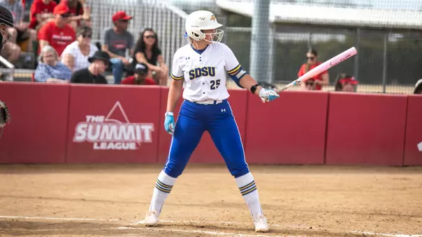 A woman in a softball uniform with a white jersey and blue pants holds a bat in her left hand while facing the pitcher as she awaits a pitch